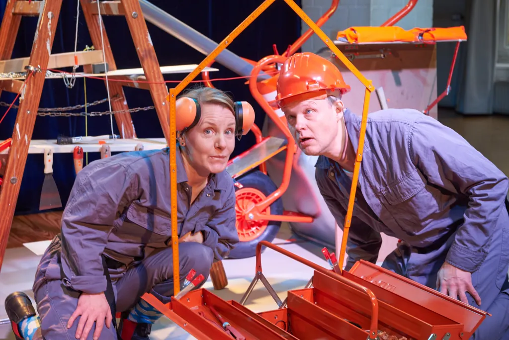Zwei Menschen in Arbeitskleidung mit Baustellenhelmen hocken in einer Baustellen-Kulisse
